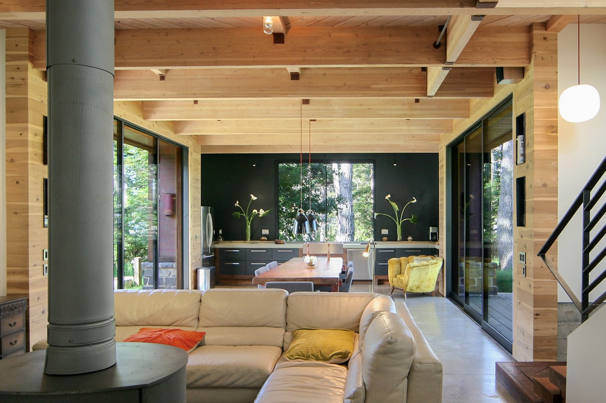 An open-concept living area features high, open beam ceilings and heated concrete floors. Large glass doors facilitate natural light and views of the surrounding forest. A cozy seating arrangement is paired with a striking wood-burning stove, creating a modern yet inviting space. A dining table sits prominently in the background.