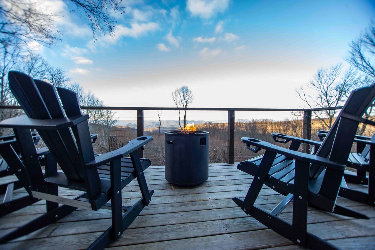 A spacious outdoor deck is furnished with several black Adirondack chairs arranged around a circular fire pit. The surrounding landscape features bare trees and open fields, with distant views under a bright sky.