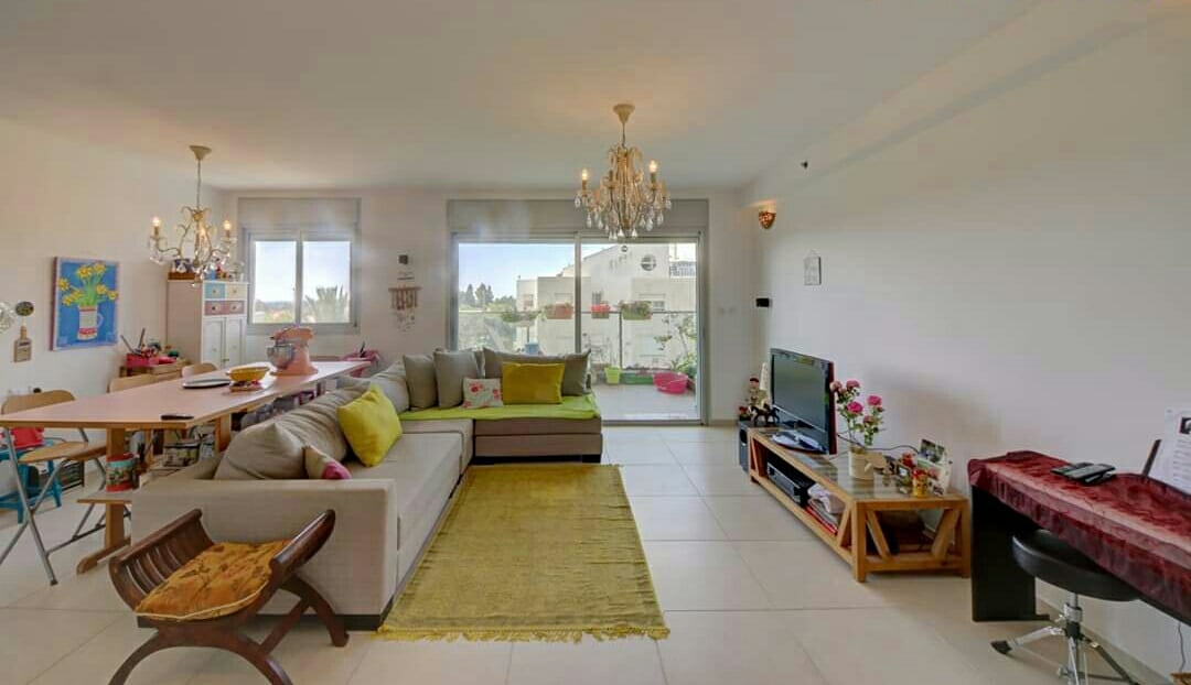 A spacious living area is brightened by natural light streaming through large windows. A comfortable sectional sofa is complemented by colorful cushions. A dining table sits nearby, adorned with decorative items, while a soft rug adds warmth to the room. Elegant light fixtures hang from the ceiling.