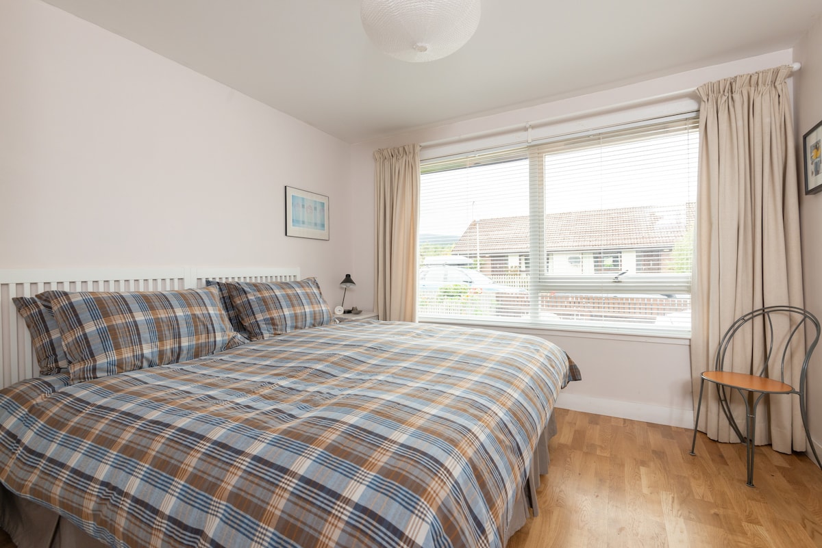 A well-lit bedroom is presented with a spacious bed adorned in a plaid pattern. Natural light fills the room through a large window, highlighting light-colored walls and wooden flooring. A bedside lamp and a simple chair add functionality to the space.