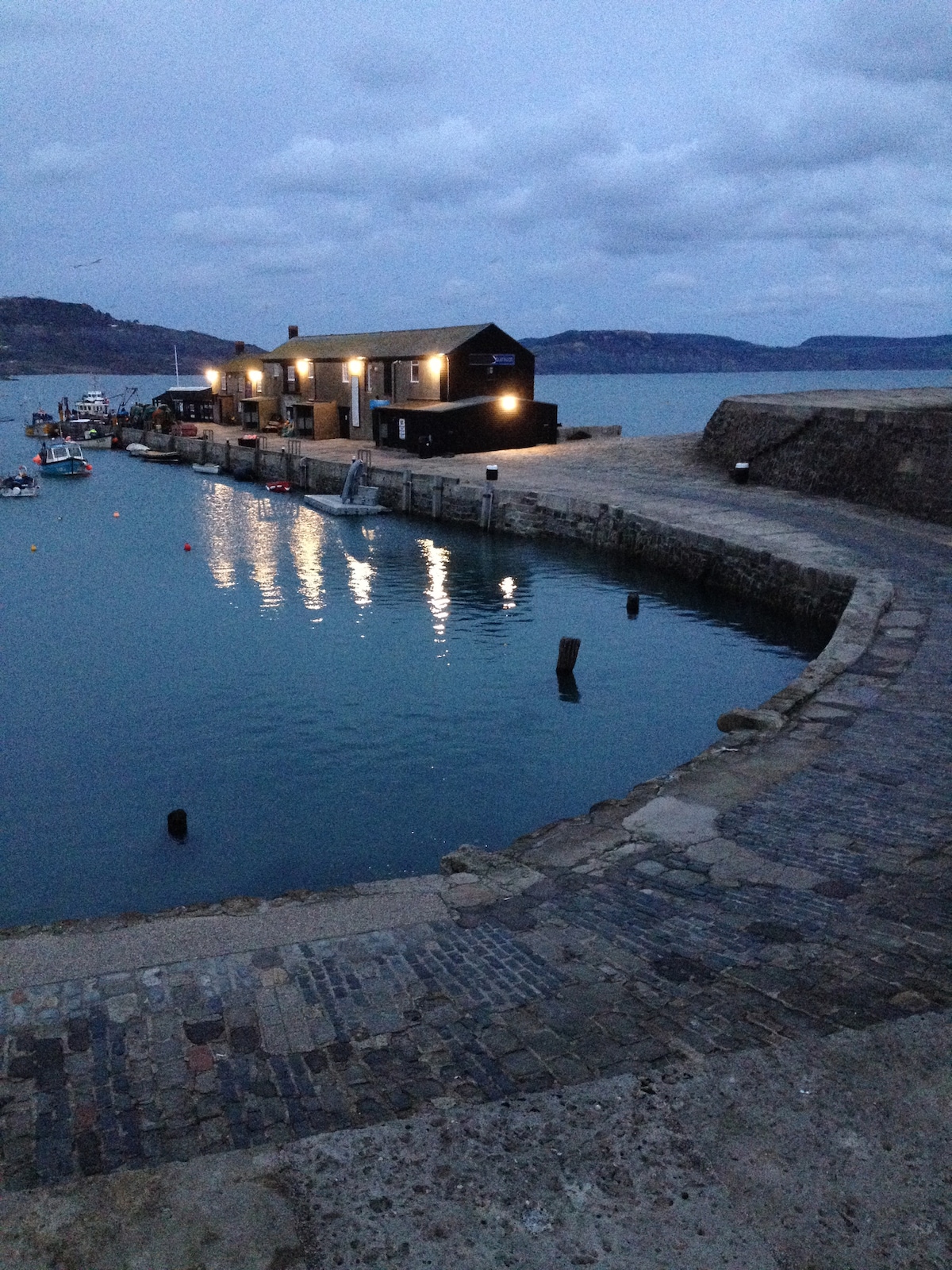 The image captures a tranquil scene of a harbor at dusk, featuring gentle waves reflecting soft light from buildings along the shore. Boats are moored peacefully, and a winding stone walkway leads to the water's edge, framed by distant hills beneath a cloudy sky.