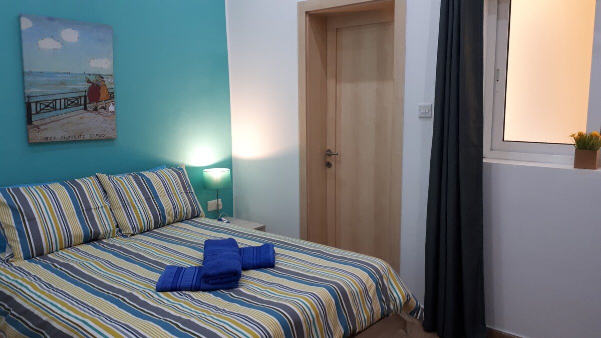 A cozy bedroom features a striped bedspread on a spacious bed, complemented by matching accent pillows. A bedside lamp casts soft light, while a small window is adorned with a dark curtain. A wooden door leads to the adjoining space.