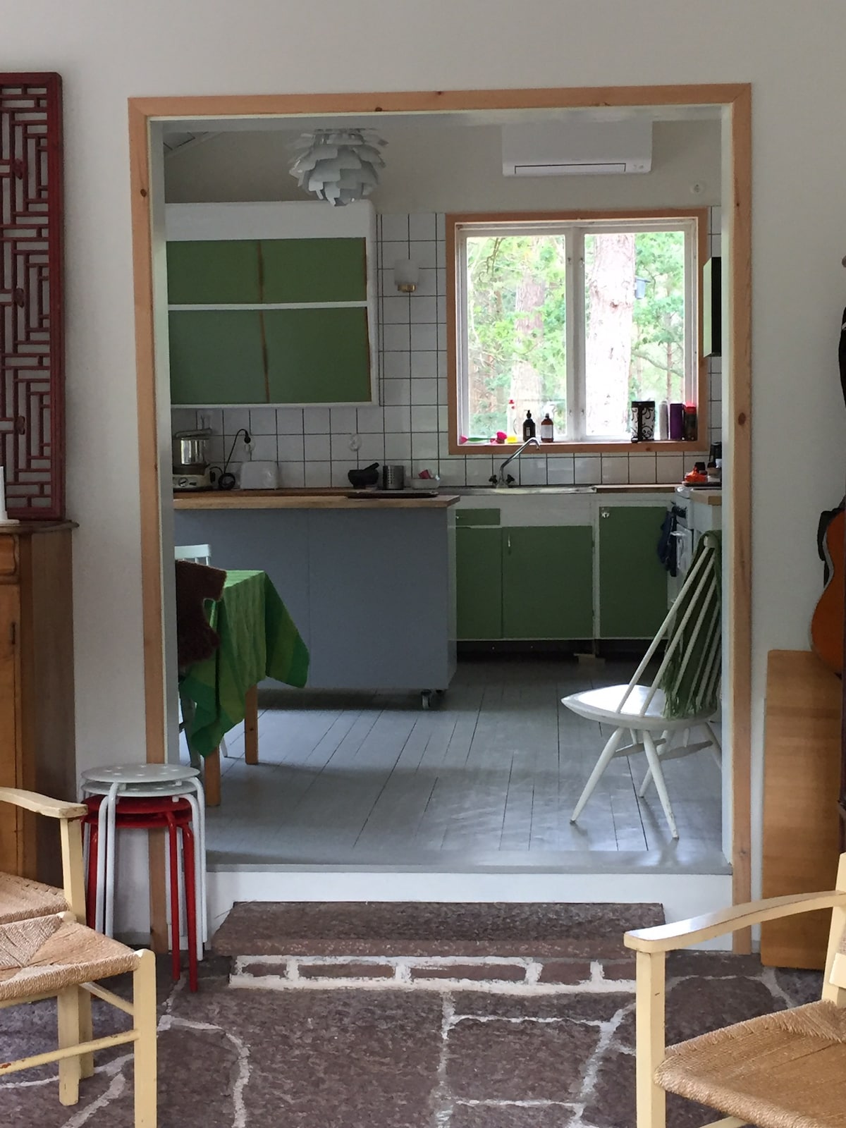 A view through a framed doorway reveals a kitchen with green upper cabinets and a contrasting backsplash. A wooden island is visible, accompanied by various kitchen appliances. Natural light enters through a window, illuminating the flooring and surrounding greenery.