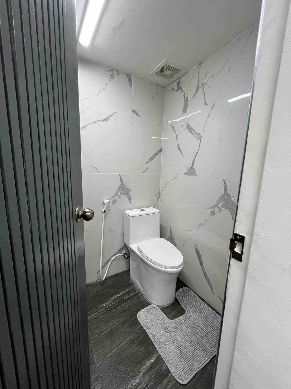 A modern bathroom features sleek marble-patterned walls and a minimalist design. A white toilet is positioned against the wall, complemented by a gray bath mat on the dark flooring. Natural light illuminates the space through a glass partition.