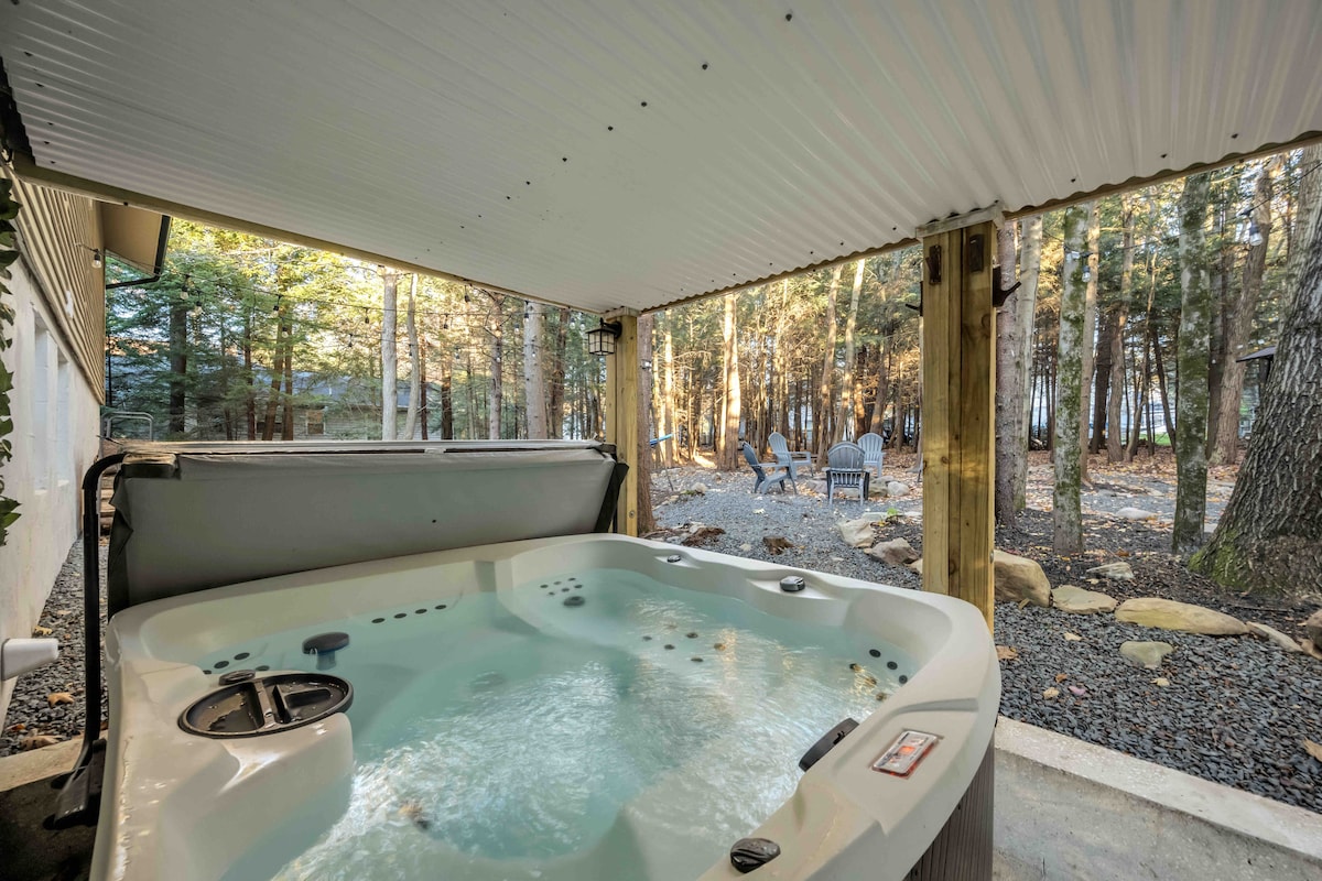 A hot tub is positioned under an awning, surrounded by wooded areas. The water is gently bubbling, with peaceful natural surroundings visible through an open space beyond. Comfortable seating is suggested by outdoor chairs in the background, enhancing the outdoor relaxation experience.