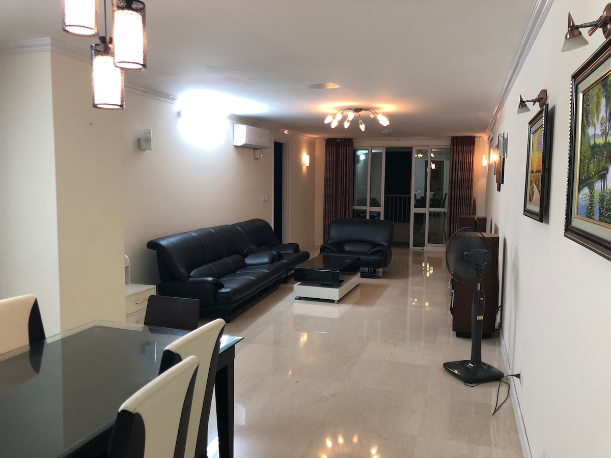A spacious living area is displayed, featuring a dark leather sofa set and a glass coffee table. Natural light pours in through large windows, highlighting the polished marble floor. Wall art is present, adding color to the neutral-toned walls. A dining table for six is visible, with modern lighting fixtures above.