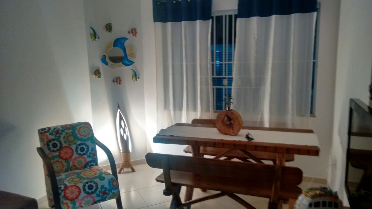 A dining area features a wooden table complemented by matching benches, set against a bright backdrop. A patterned chair adds color, while decorative fish adorn the wall. Natural light filters through sheer curtains, enhancing the inviting atmosphere of the space.