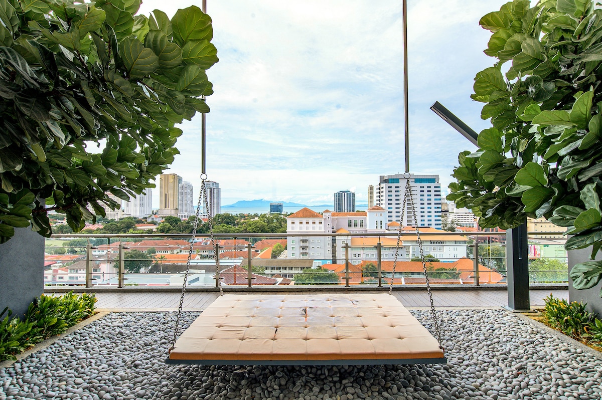 A spacious swing seat is surrounded by lush greenery, suspended above a textured stone surface. The view showcases a blend of city buildings and distant hills under a clear sky.