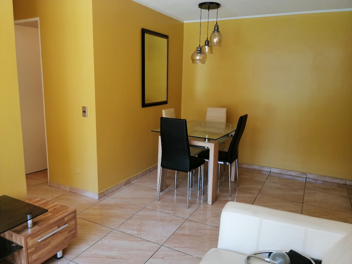 A bright dining area features a glass table surrounded by four black chairs under pendant lighting. A large mirror is positioned on the wall, reflecting the warm yellow hue that accents the space. The tiled floor adds a modern touch.