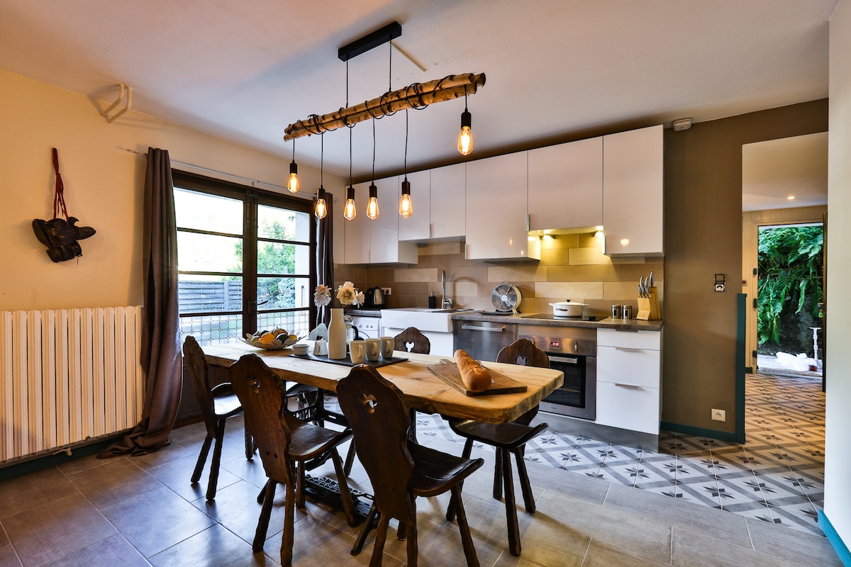 The kitchen features modern appliances and a spacious dining table with wooden chairs. Natural light enters through large windows, highlighting the clean lines and warm tones of the cabinetry. Decorative elements include a rustic light fixture and subtle wall decor, contributing to a welcoming ambiance.