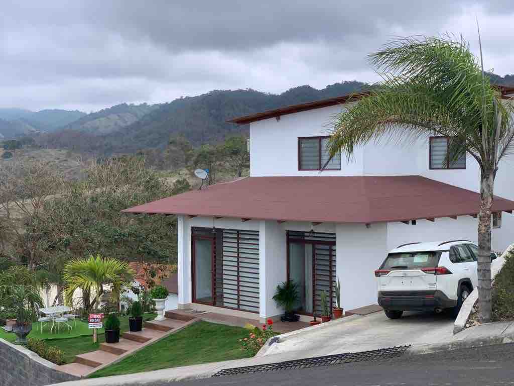 A contemporary two-story house with a sloped roof is situated on a hillside. Large windows provide natural light. A vehicle is parked on the driveway. Lush greenery and a few potted plants enhance the surroundings, while distant mountains create a scenic backdrop.