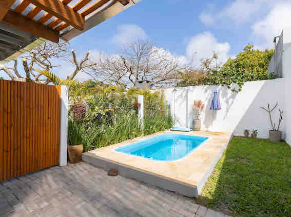 A private outdoor area is featured, showcasing a small swimming pool surrounded by well-maintained greenery and stone pathways. A wooden gate offers access, while a sunny sky and minimalistic decor enhance the tranquil ambiance.