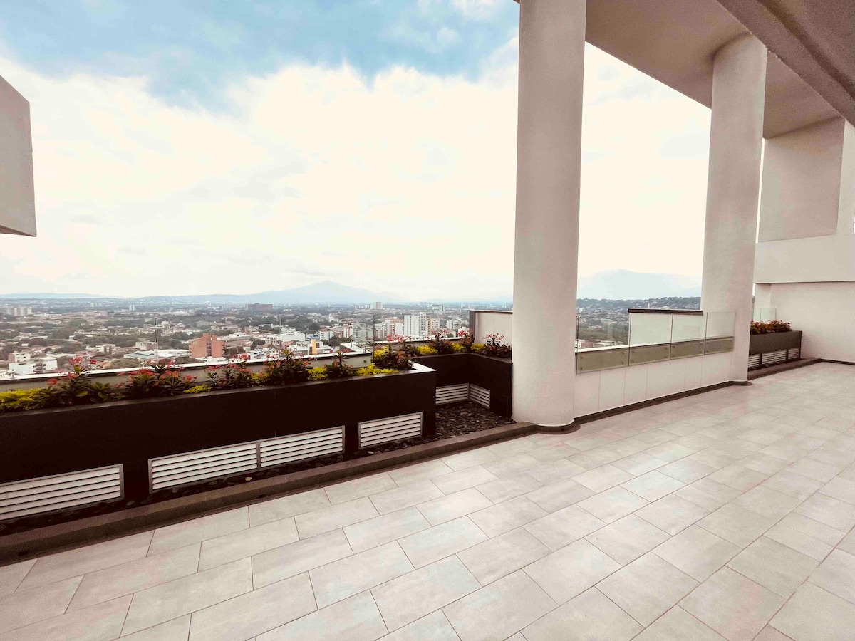 A spacious terrace is presented with a smooth tiled floor and large open spaces. Lush planters line the edges, showcasing various plants. The scenic view of the surrounding landscape, featuring mountains and city buildings, can be observed in the distance under a cloudy sky.