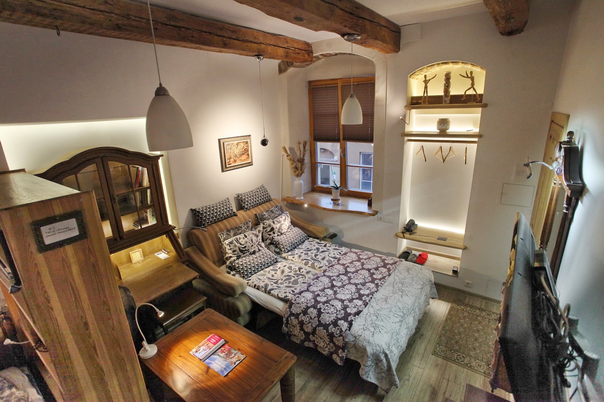 A spacious room features exposed wooden beams, creating a rustic ambiance. A large bed dressed in dark patterned bedding is positioned between two windows. A wooden table is visible, with a local magazine casually placed on its surface. Decorative items are displayed on a shelf along the wall.
