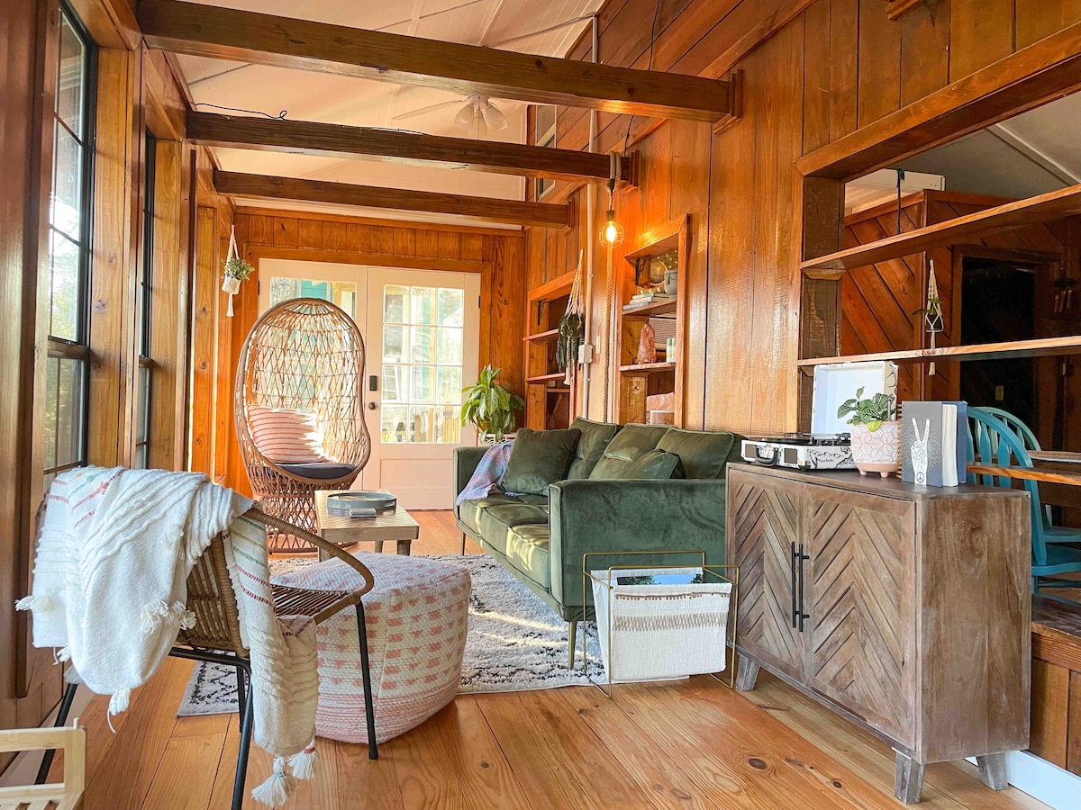 The living area features warm wooden paneling and a cozy seating arrangement. A green sofa and colorful accent chairs are present, along with a light-filled entryway. Decorative shelving lines the walls, complemented by a woven chair and a patterned area rug.