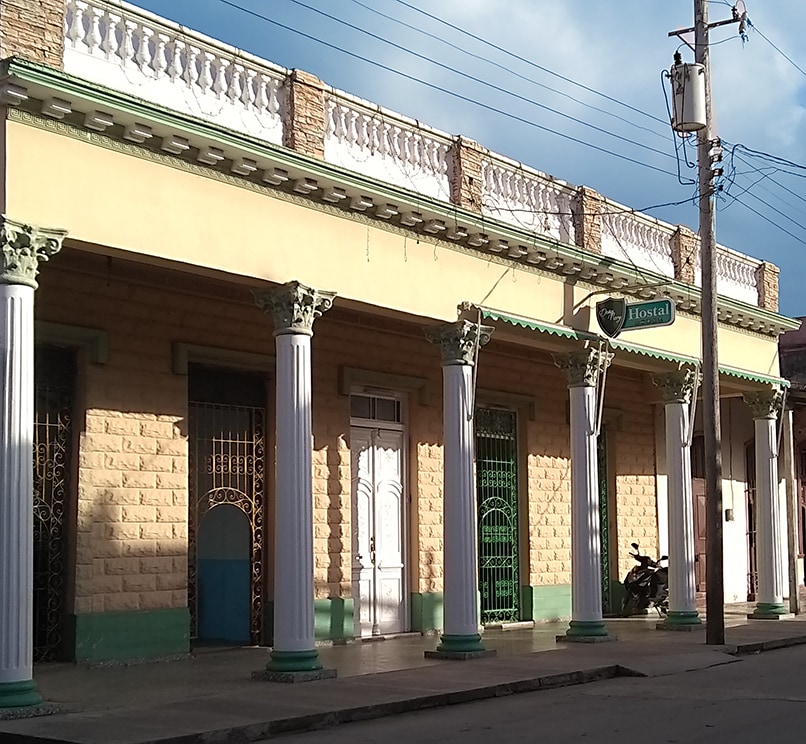 The colonial-style facade of the Hostal Doña Nery features tall columns and decorative stonework. Green accents enhance the exterior, and large windows allow natural light to enter. A motorcycle is parked nearby, while the building's intricate door designs add character to the entrance.