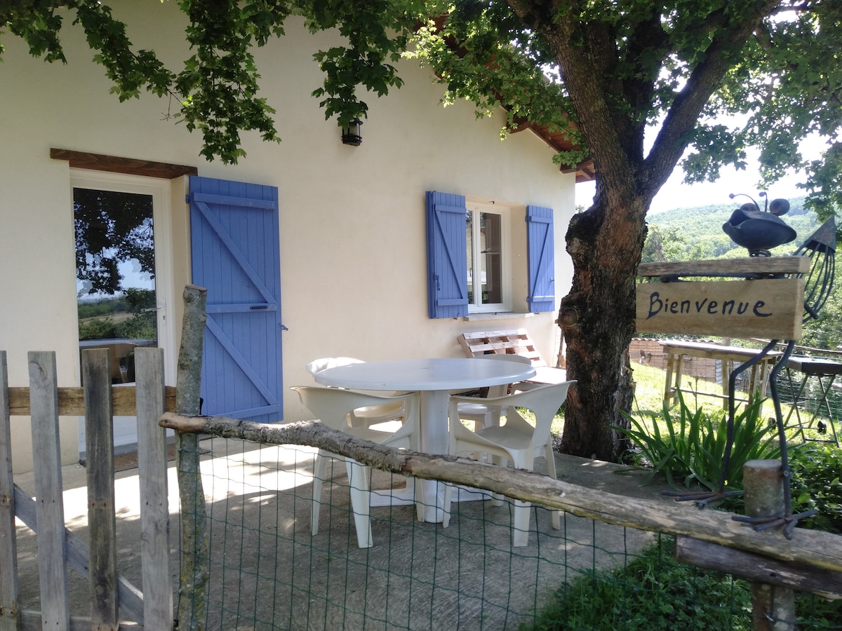 A welcoming exterior view of the gîte features light blue shutters against a neutral wall. A round table is set on a stone patio, accompanied by wooden seating. A tree shades the area, enhancing the natural setting. A wooden sign reads 'Bienvenue.'