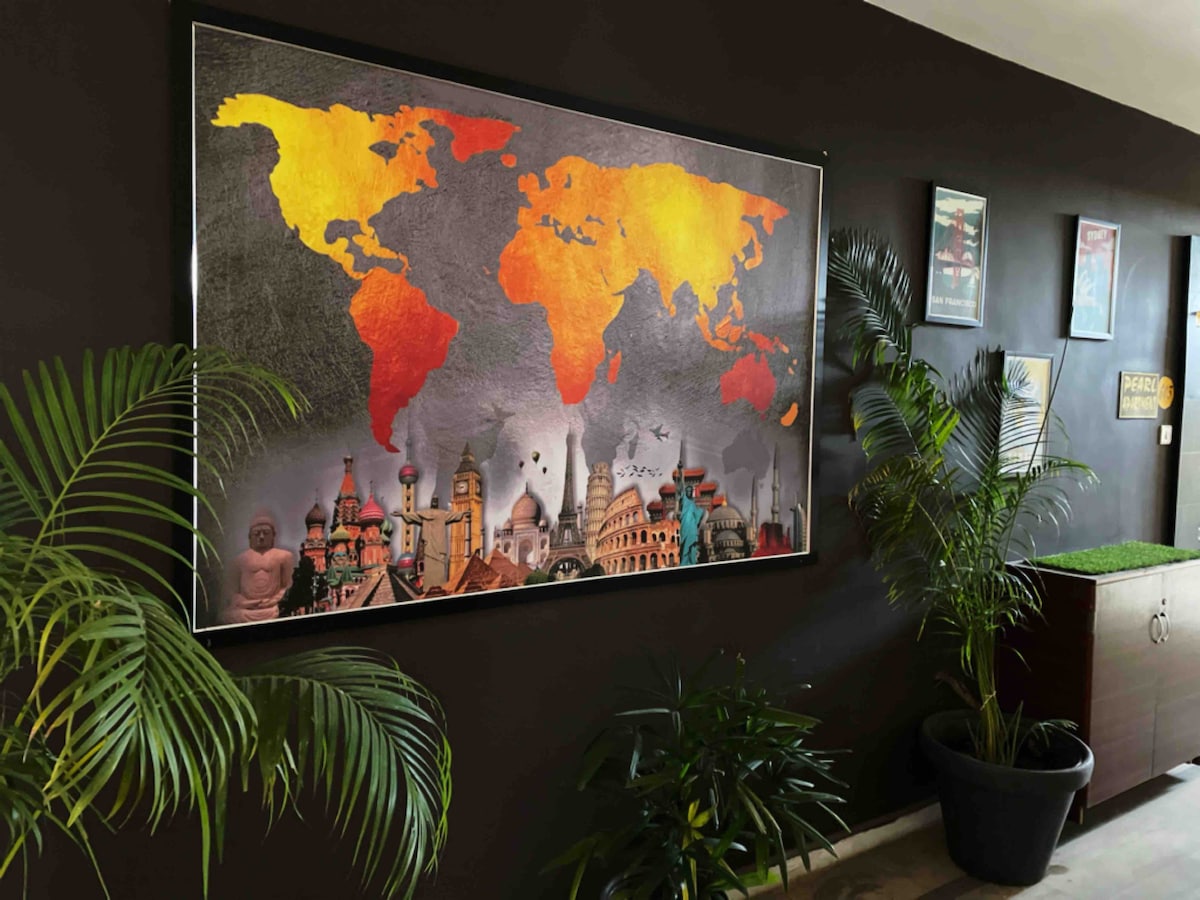 A large framed world map adorns a dark wall, featuring vibrant shades of orange and yellow. The map is complemented by a collage of iconic landmarks from various countries. Potted plants are placed nearby, enhancing the decor of the corridor.