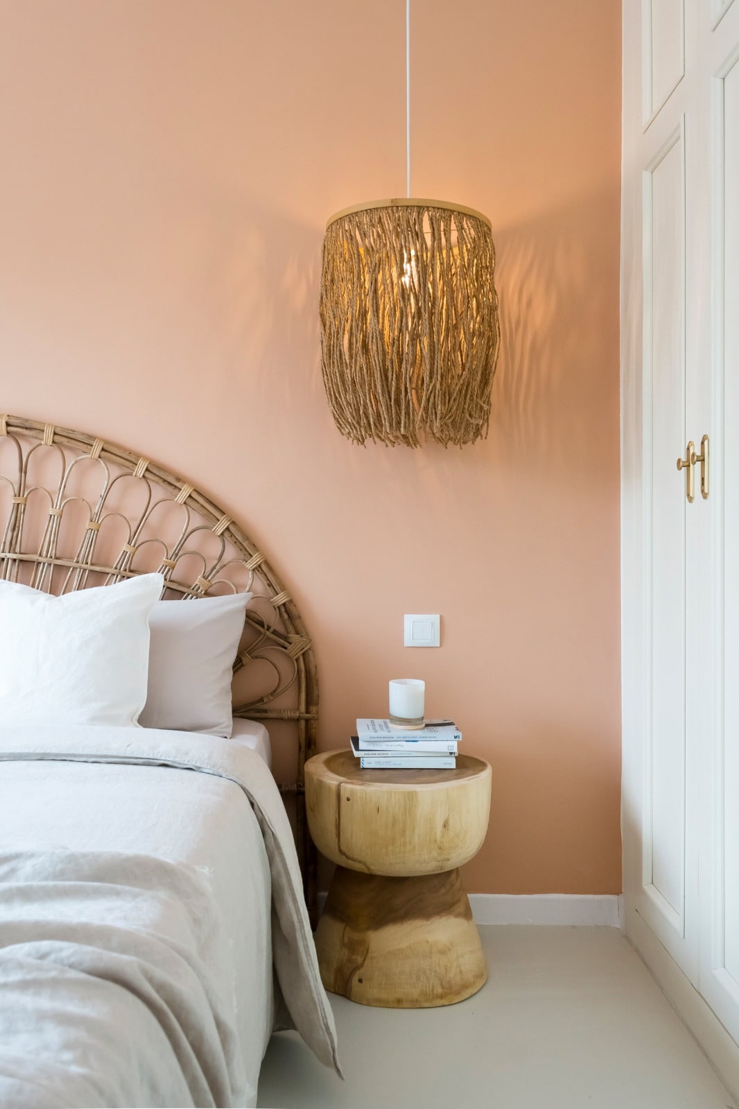A softly lit bedroom features a natural wood headboard against a peach-colored wall. A small, round wooden side table is placed beside the spacious bed, holding a stack of magazines and a candle. A light fixture made of woven material hangs above, enhancing the room's cozy ambiance.