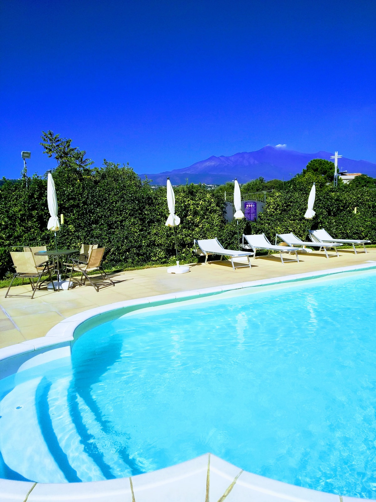 A refreshing swimming pool is outlined by a white deck, featuring several lounge chairs and small tables with umbrellas. Lush green foliage surrounds the area, while a mountain is visible in the background against a clear blue sky.