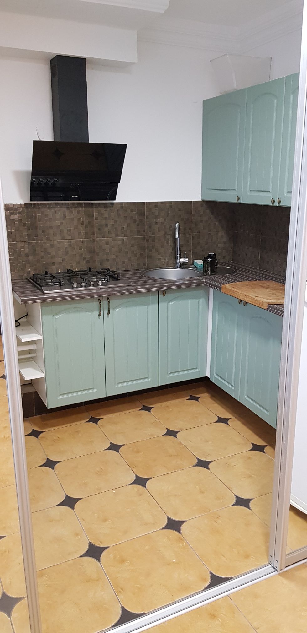 A modern kitchen area is characterized by mint green cabinetry and a gas stove with an exhaust hood. A stainless steel sink is positioned beneath the cabinetry, with a countertop surface adjacent to a wooden cutting board. The tiled floor features a geometric pattern of light and dark colors.