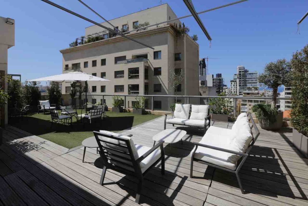 A spacious rooftop terrace features multiple seating areas with comfortable white cushions. The wooden deck is complemented by greenery and a large umbrella for shade, creating an inviting space for relaxation. Views of surrounding buildings and cityscape are visible in the background.