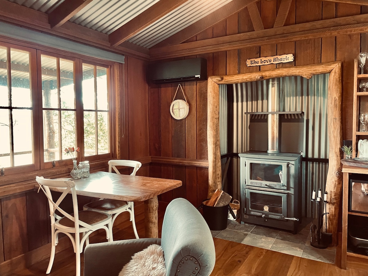 A wooden interior showcases a cozy seating area with a light blue chair and a rustic wooden table. A wood-burning stove is positioned against a corrugated backdrop, with a decorative sign above it. Natural light floods in through multiple windows, enhancing the warmth of the space.