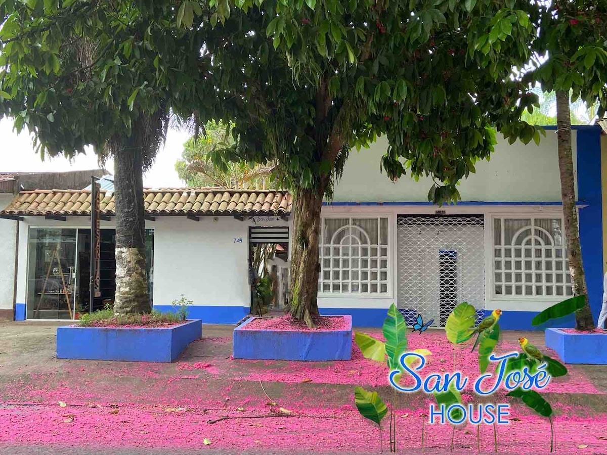 A charming house features a bright blue exterior complemented by a white facade. Lush greenery surrounds the property, with a large tree providing shade. The ground is covered with vibrant pink petals, creating a colorful contrast against the earth-toned path.