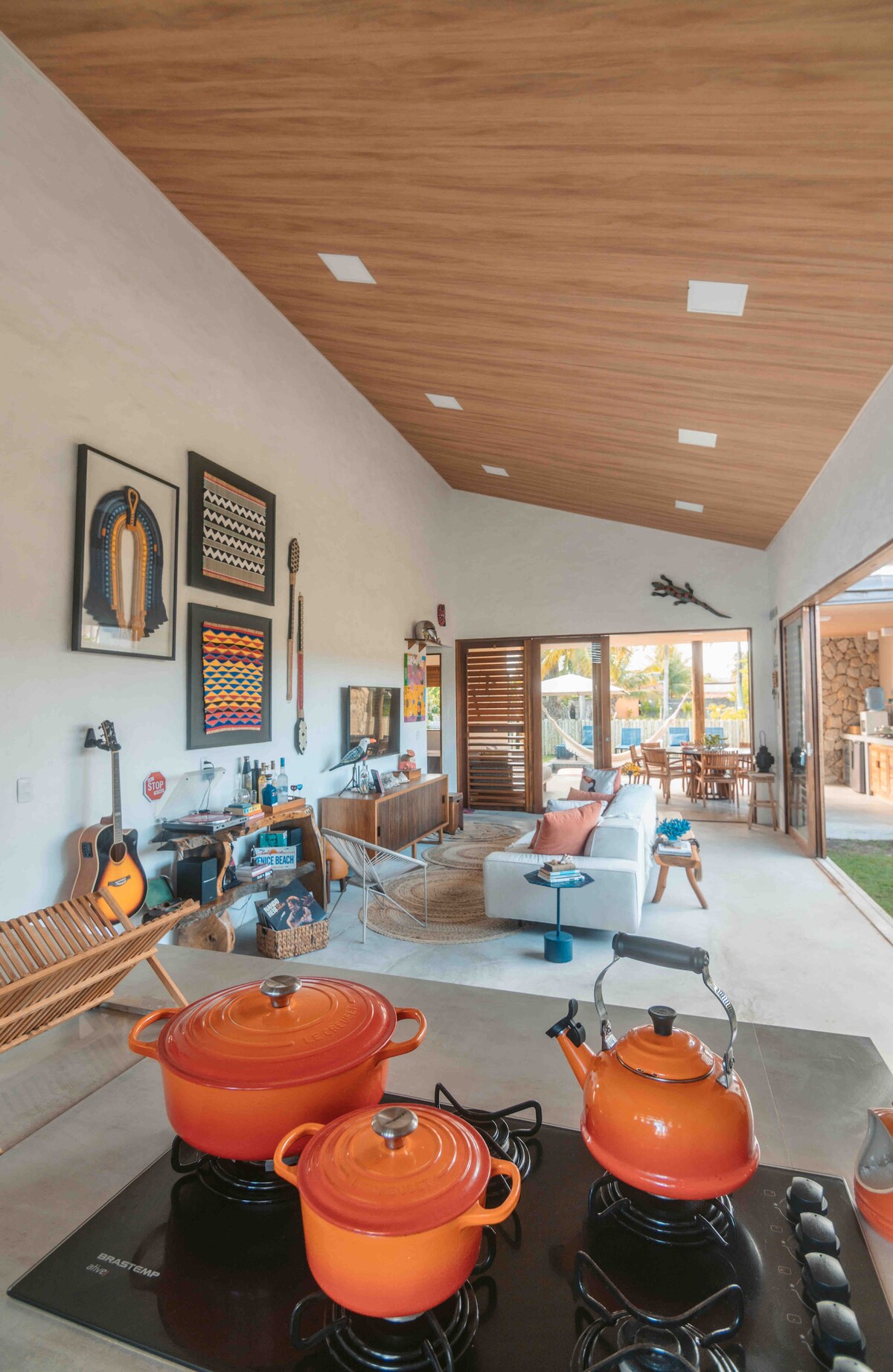 A modern living space is highlighted, featuring a high wooden ceiling and large glass doors that open to the outdoors. Vibrant orange cookware is neatly arranged on a gas stove, while a comfortable seating area is visible in the background, complemented by artistic wall decorations.