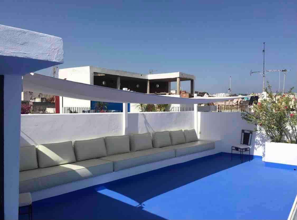A spacious rooftop terrace features a built-in seating area with soft cushions, designed for relaxation. The area is surrounded by a white wall and has a vibrant blue floor, creating a bright and refreshing space with clear skies above.