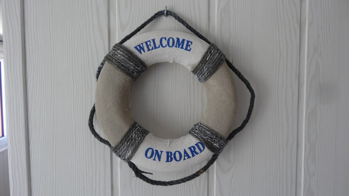 A nautical-themed decorative life preserver hangs on a wooden wall. The life preserver displays the text 'WELCOME ON BOARD' in blue lettering, providing a friendly and inviting touch to the space.