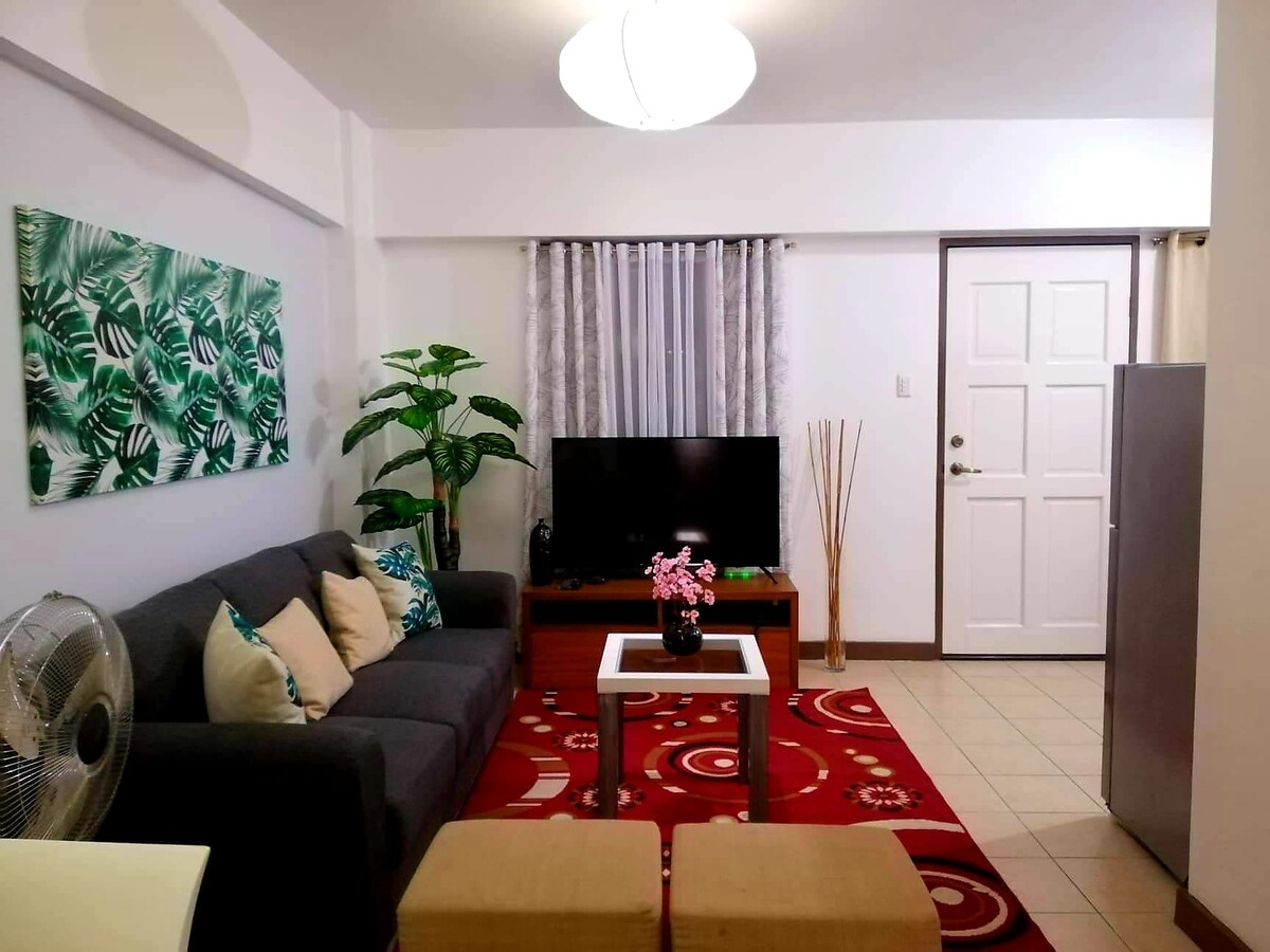 A cozy living area is highlighted by a grey sofa adorned with decorative cushions. A coffee table sits in front of the sofa, complementing a vibrant red area rug. A television is positioned against the wall, surrounded by indoor plants, under soft ambient lighting.