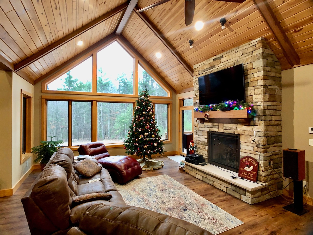 A spacious living area features high vaulted ceilings with wooden beams and large windows that provide natural light. A stone fireplace is centered along one wall, complemented by a decorated Christmas tree. Comfortable seating arrangements include a large brown sectional couch and a plush chaise lounge.