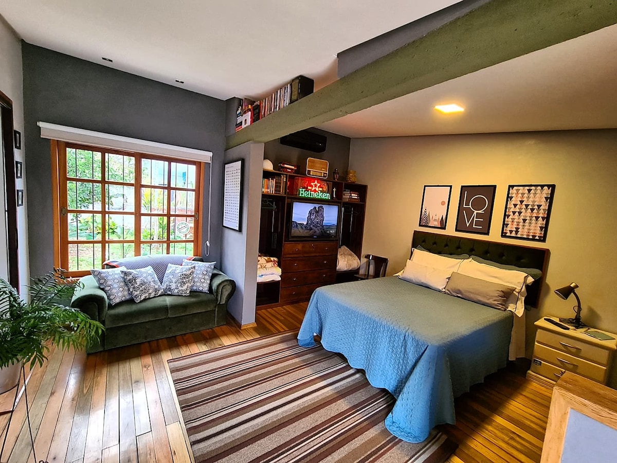A cozy sleeping area features a spacious bed draped with a light blue coverlet. A green sofa complements the warm wooden flooring, and natural light streams through a large window, creating an inviting ambiance. Bookshelves are visible along the wall, adding character to the room.
