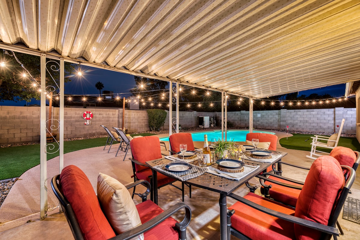 An outdoor dining area is situated beneath a covered patio, featuring a table set with plates and drinks. Surrounding comfortable red chairs invite relaxation, while string lights add soft illumination. In the background, a heated swimming pool and a putting green are visible, creating a tranquil setting.