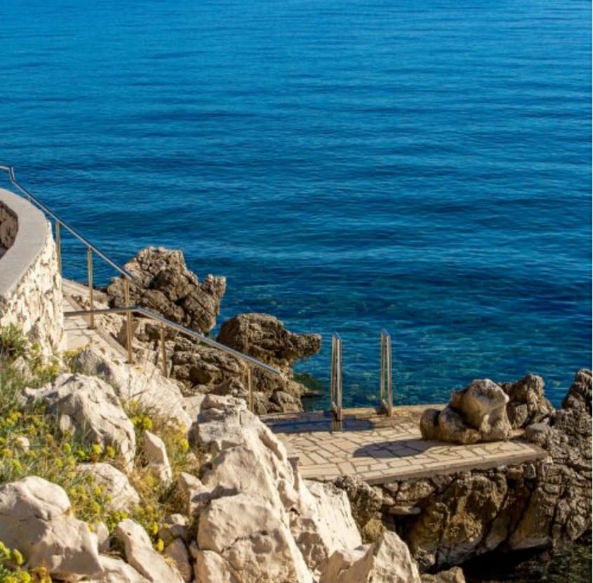 A rocky shoreline is depicted with a gently sloping pathway leading to the water's edge. Steps are visible, providing access to the clear, blue water. Surrounding greenery adds a natural element to the coastal landscape.