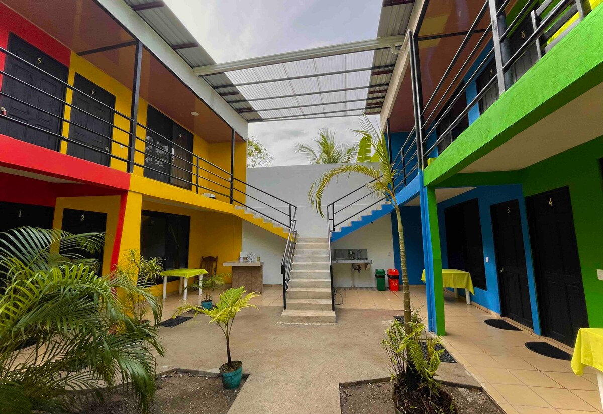 The image showcases a vibrant courtyard surrounded by colorful cabins, with black doors numbered from 1 to 5. A staircase leads to the upper level, and light yellow tables are positioned on the ground level. Lush greenery enhances the inviting atmosphere.
