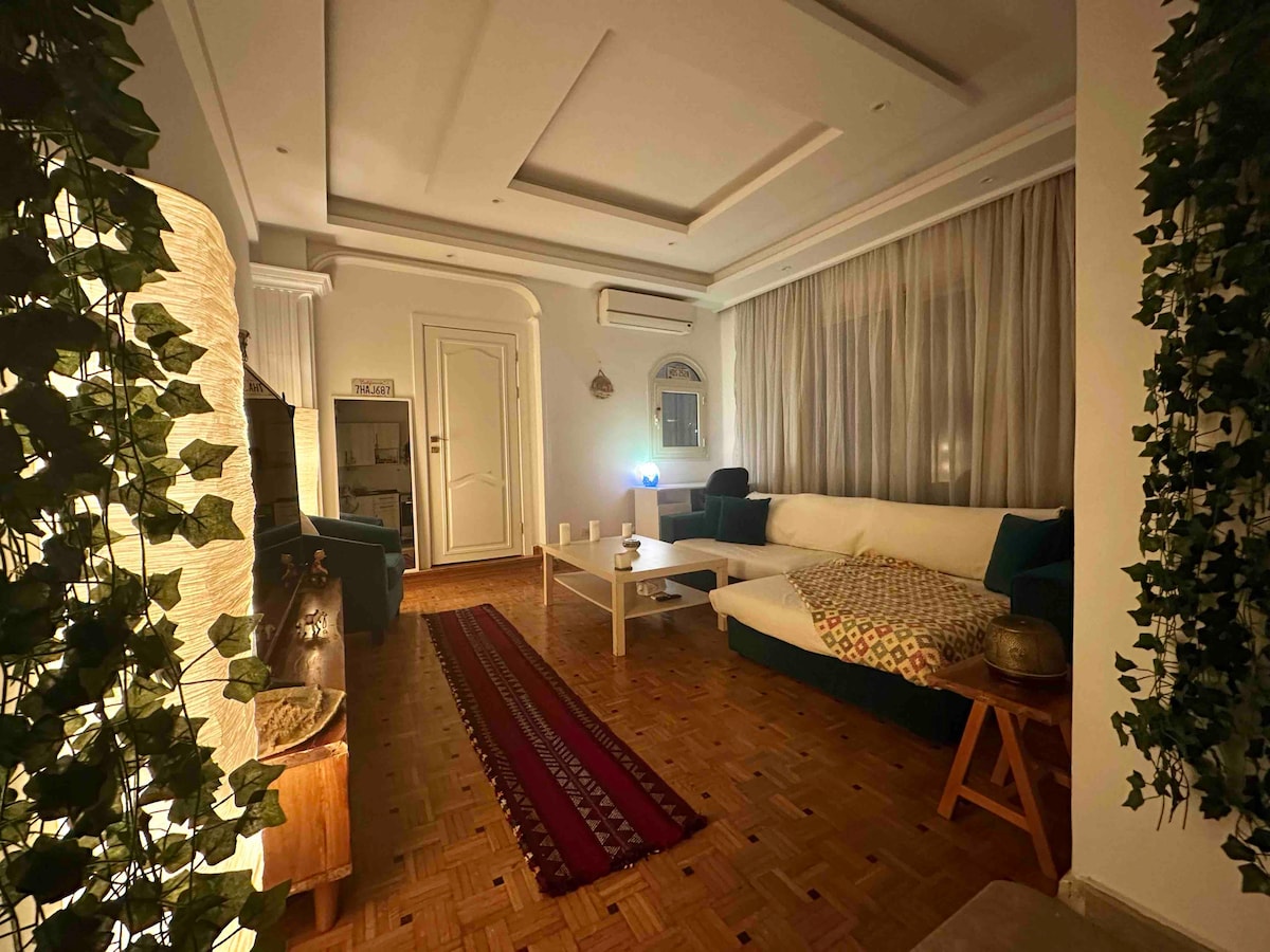 A spacious living area is illuminated by soft lighting, creating a calm ambiance. A comfortable sofa is arranged around a low table, with a patterned rug anchoring the space. Large windows are dressed with sheer curtains, allowing filtered light to enhance the inviting decor.