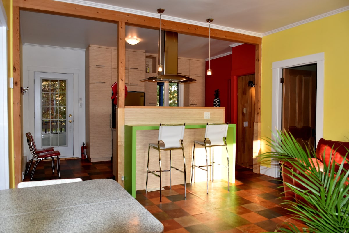 A stylish kitchen area features a modern bar with two high stools and a contrasting green countertop. Light pours in through windows, highlighting the warm tones of the wooden beams and colorful walls, creating an inviting space for gatherings.