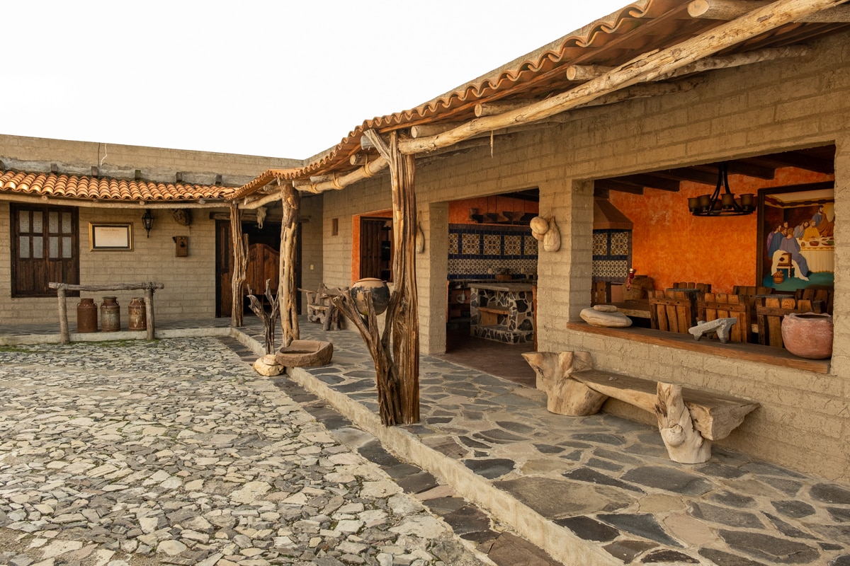 An inviting outdoor area features a stone-paved pathway leading to a spacious dining area. Large windows offer views of rustic wooden beams and colorful murals. Natural textures and earthy tones are present in the furniture and decor, creating a rustic yet comfortable environment.