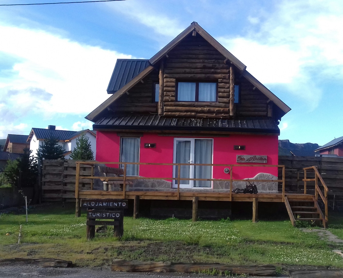 A charming cabin with a vibrant pink facade and wooden accents is featured. A spacious deck extends from the front, offering seating areas for relaxation. The entrance is marked by a welcoming sign, and the surrounding green lawn provides a serene outdoor space.