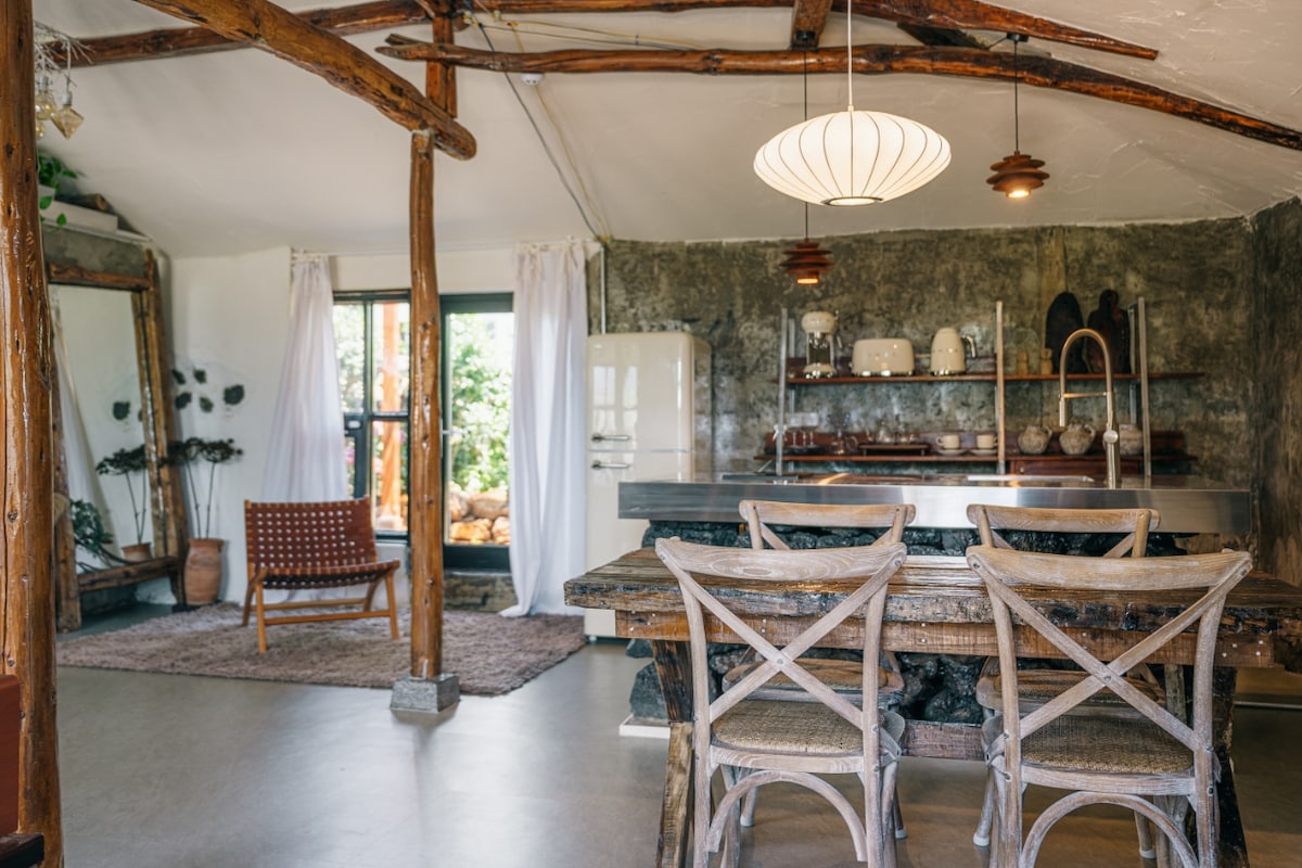 A dining area features a rustic wooden table surrounded by light-colored chairs. Exposed wooden beams enhance the ceiling. A vintage refrigerator and open shelving display dishware against a textured stone wall, while natural light enters through the large window draped with white curtains.