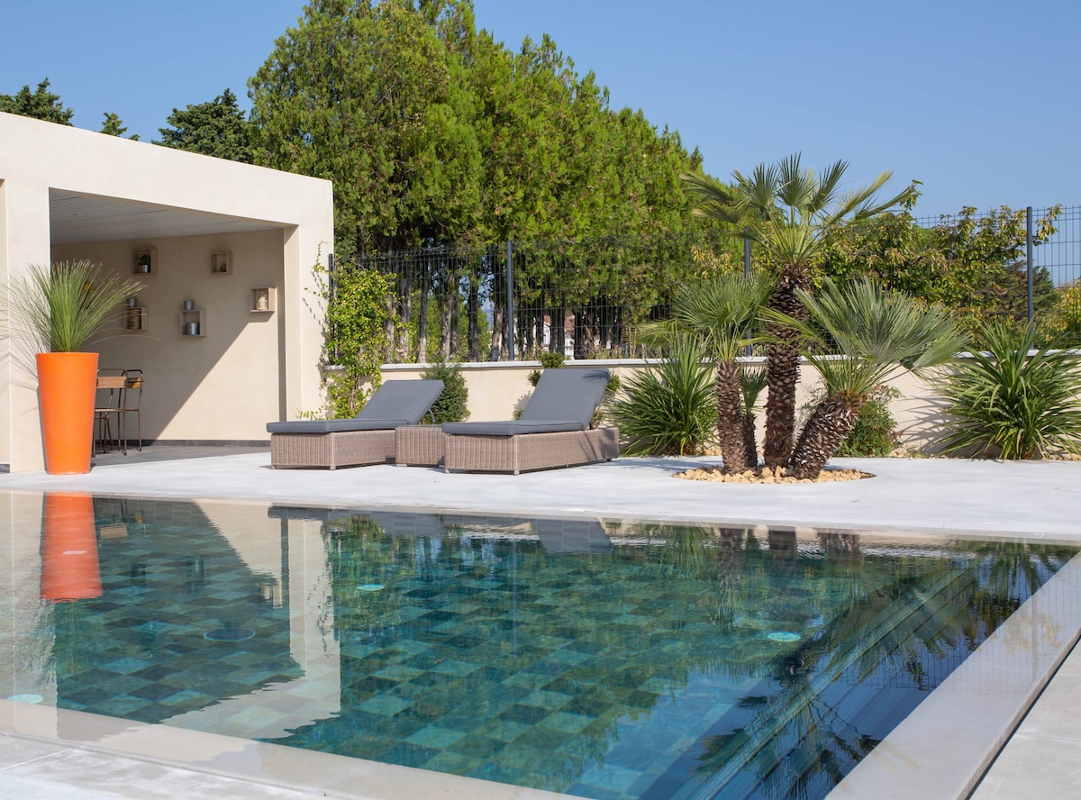 A serene outdoor space is highlighted by an elegant infinity pool that reflects the sky. Sun loungers are arranged nearby, surrounded by lush greenery and textured landscaping elements. A cozy seating area is positioned under a covered terrace, perfect for relaxation.