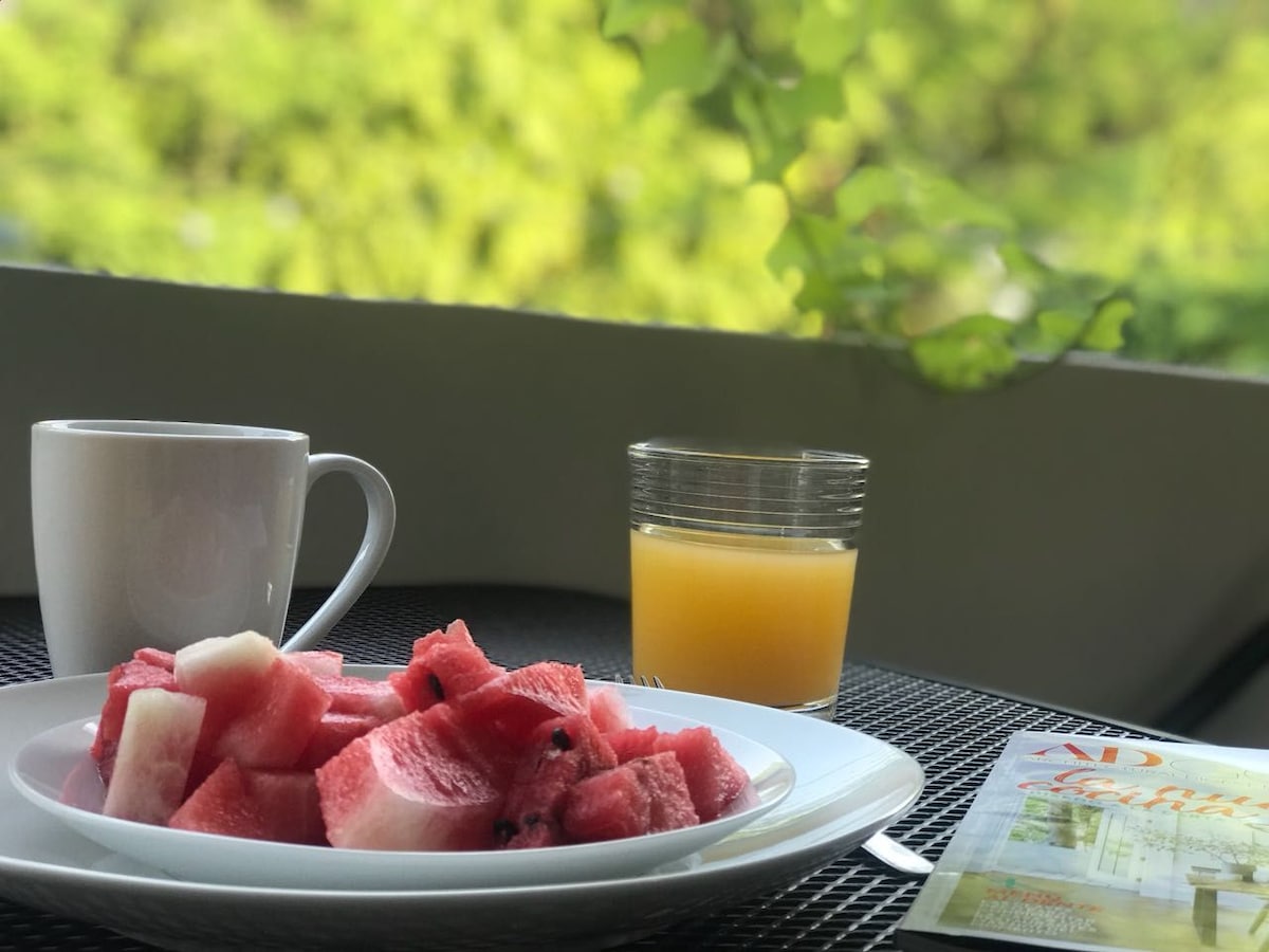 A breakfast setting is displayed on a textured table. A white plate holds neatly cut pieces of watermelon, while a cup of coffee sits beside it. A glass of orange juice reflects sunlight, and a magazine rests nearby, with a backdrop of lush greenery visible.