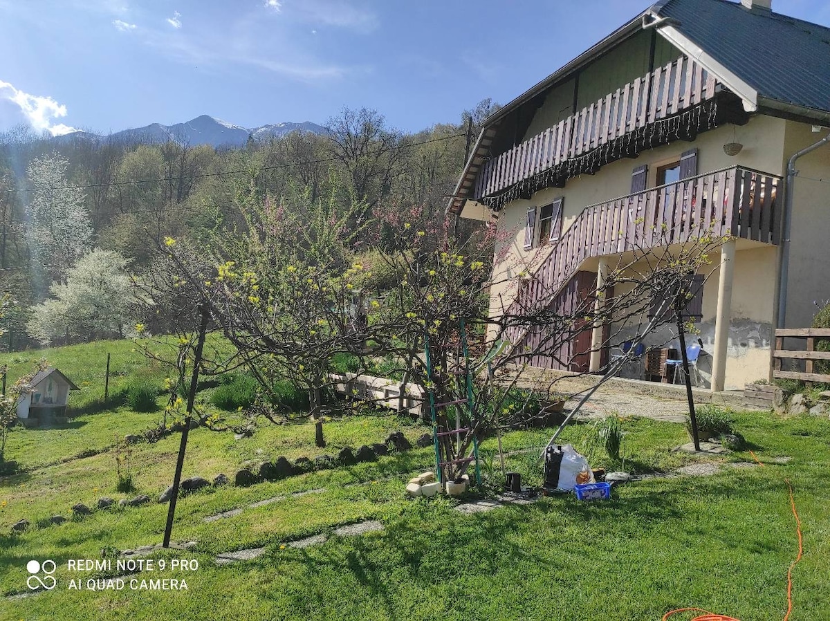 A charming exterior view of the accommodation reveals a two-story house with a sloped roof and a balcony adorned with wooden railings. The surrounding garden features a well-maintained lawn and various trees. The scenic backdrop of the mountains is visible in the distance.