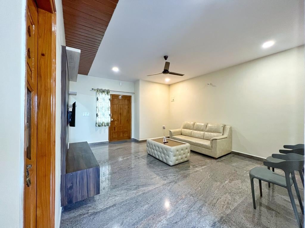 A spacious living area features a light-colored sofa and a sleek coffee table on a polished stone floor. A flat-screen television is mounted on the wall, and an elegant wooden door leads to the outside. Natural light enters through the window, framed by sheer curtains.