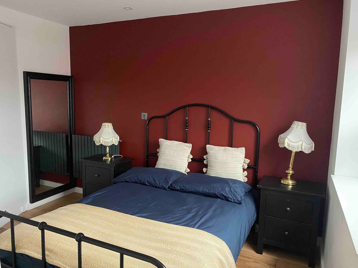 A cozy sleeping area features a metal bed frame with a blue duvet and two plush pillows. Flanking the bed, two dark nightstands hold decorative lamps. A large mirror reflects the room’s warm red wall, enhancing the inviting atmosphere.