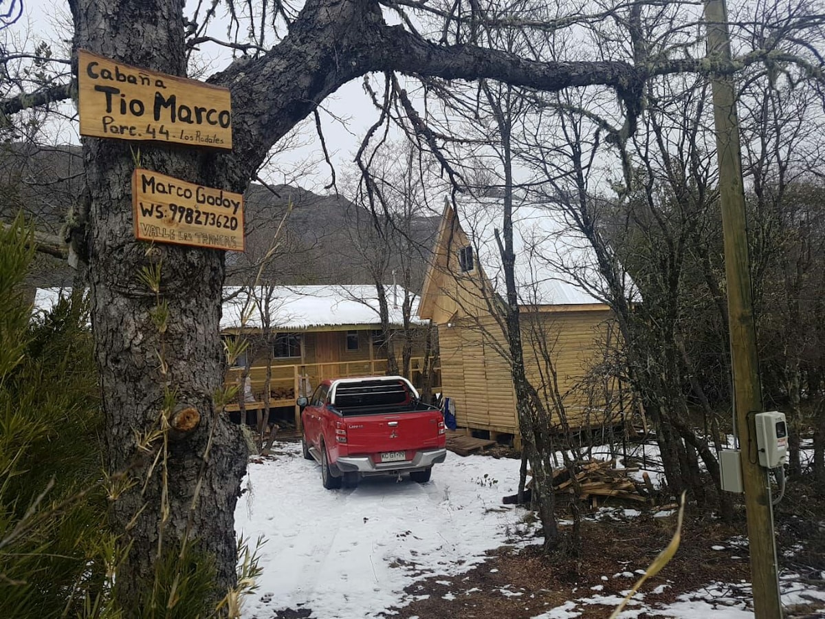 The image features the cabin named 'Cabaña Tio Marco,' nestled among trees with a snowy landscape. A red pickup truck is parked on a gravel path leading up to the wooden structure, which displays a sloped roof and large windows.