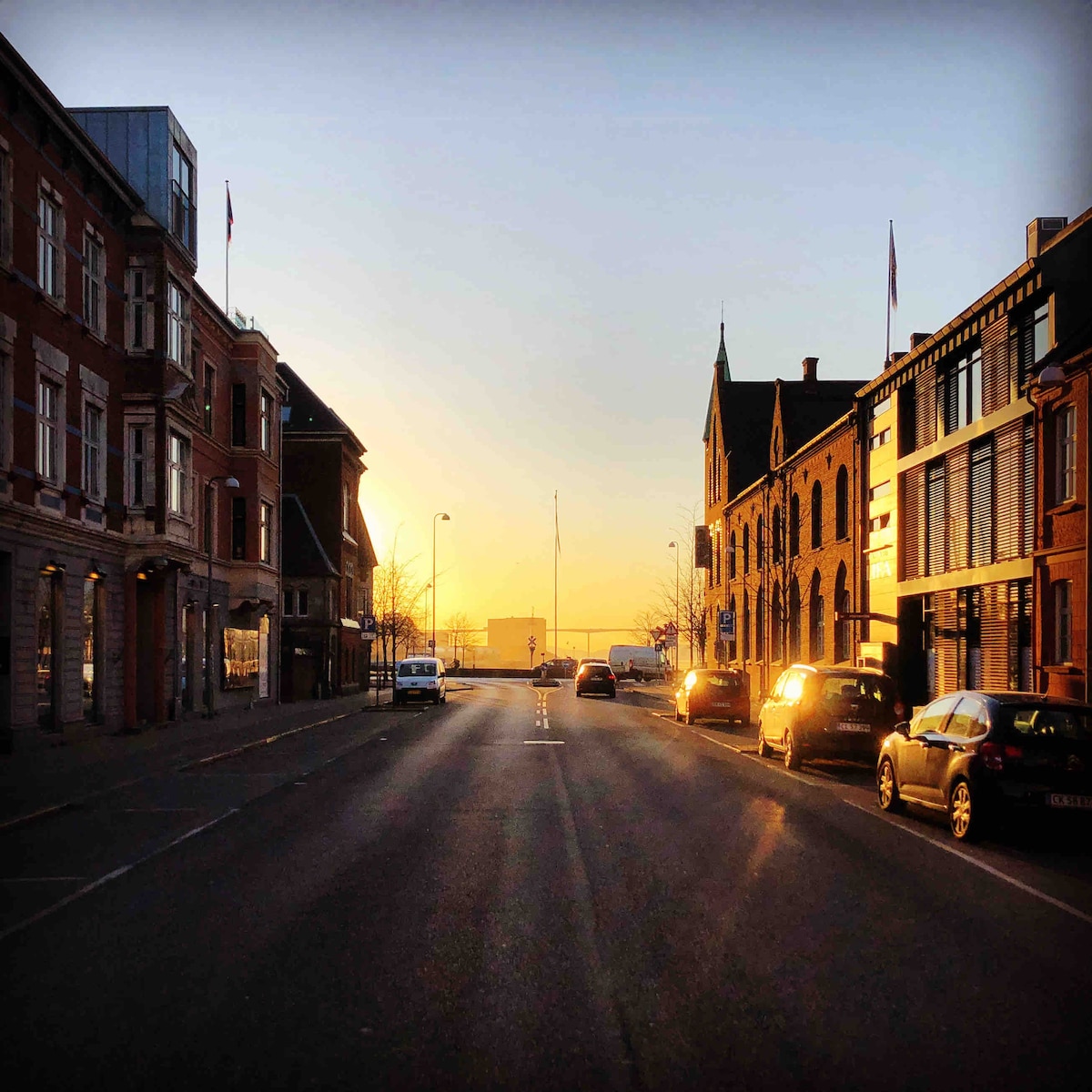 A wide street is depicted, lined with buildings on either side, showcasing a variety of architectural styles. The sun sets in the background, casting a warm glow over the scene. Cars are parked along the street, and soft light reflects off the windows.