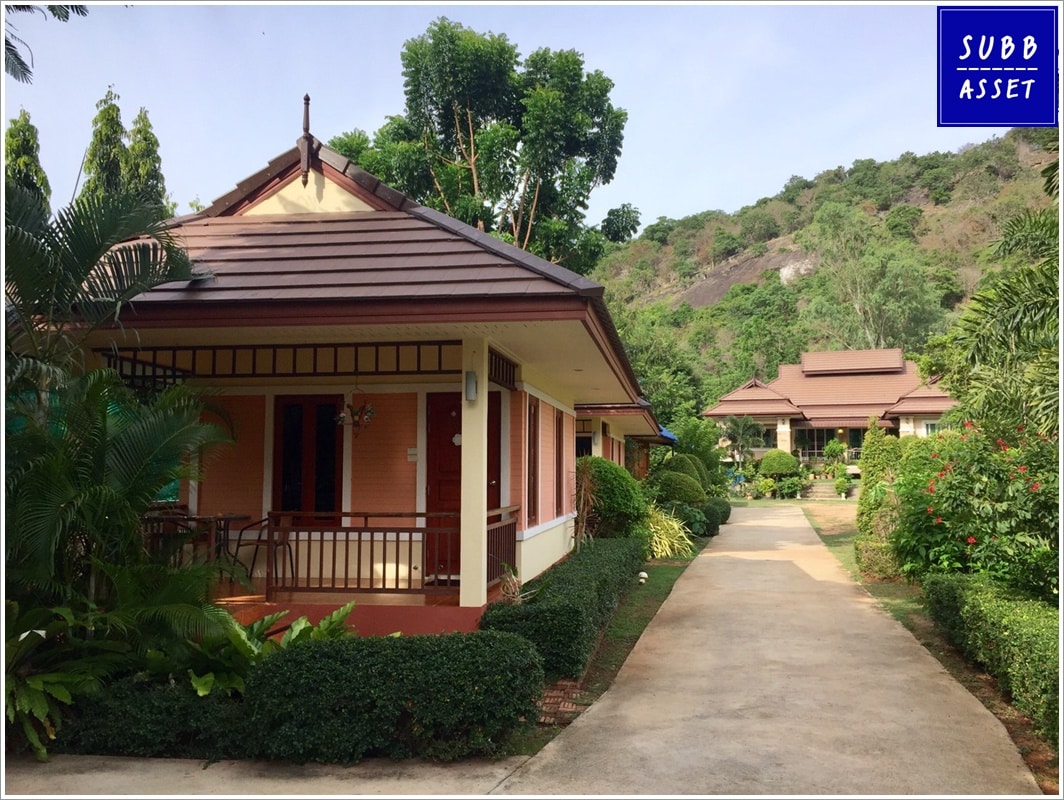 Two private houses are situated along a tidy pathway, surrounded by lush greenery and well-maintained gardens. Each house features a warm-toned exterior with a sloped roof. The landscape includes manicured hedges and tropical plants, creating a serene environment.